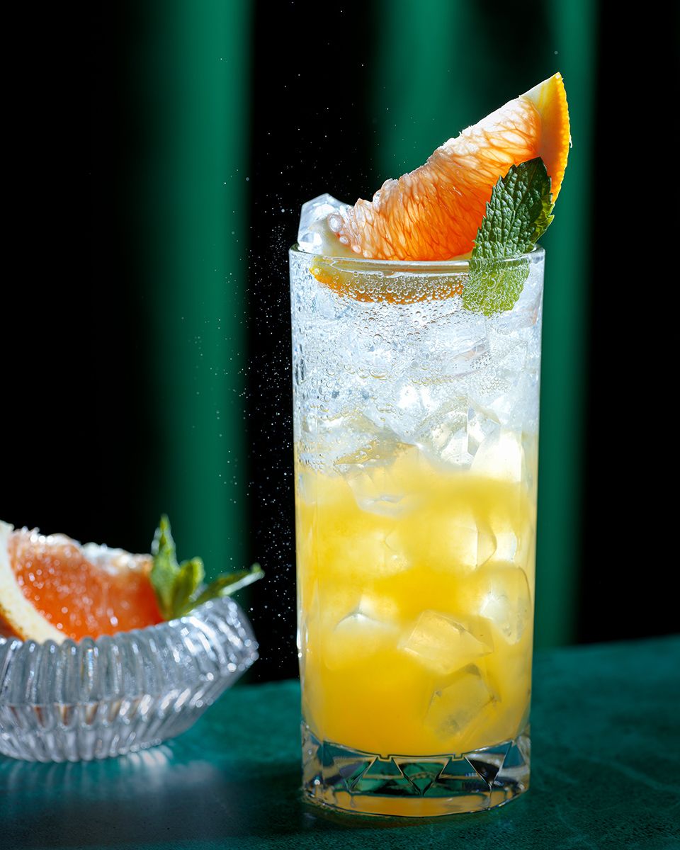 A refreshing grapefruit cocktail with ice in a tall glass, garnished with a citrus wedge and mint leaf.