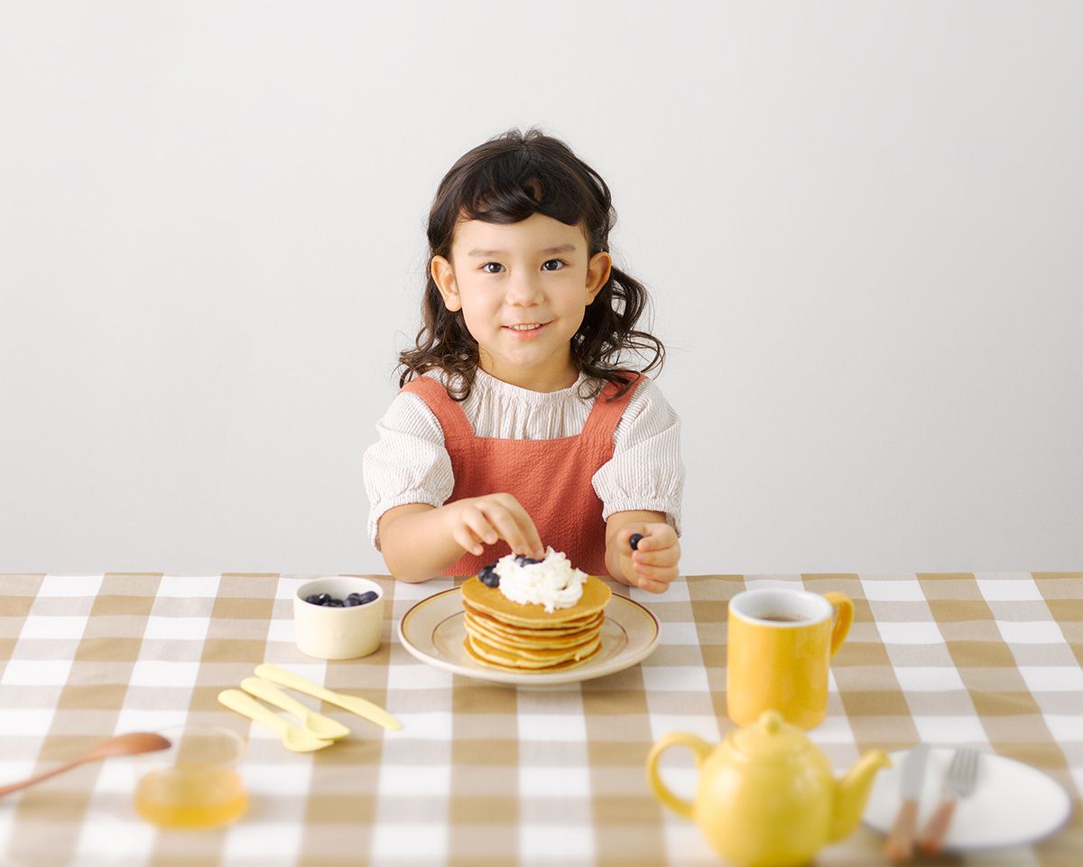 Young girl smiling at a table with a stack of pancakes topped with whipped cream on a checkered tablecloth.