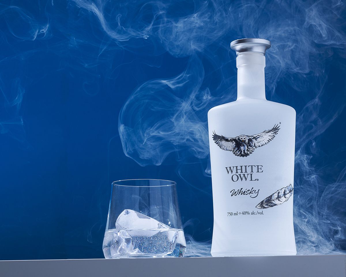 White Owl Whisky bottle in frosted white glass with an owl design, beside a rocks glass with ice, against a blue smoky background.
