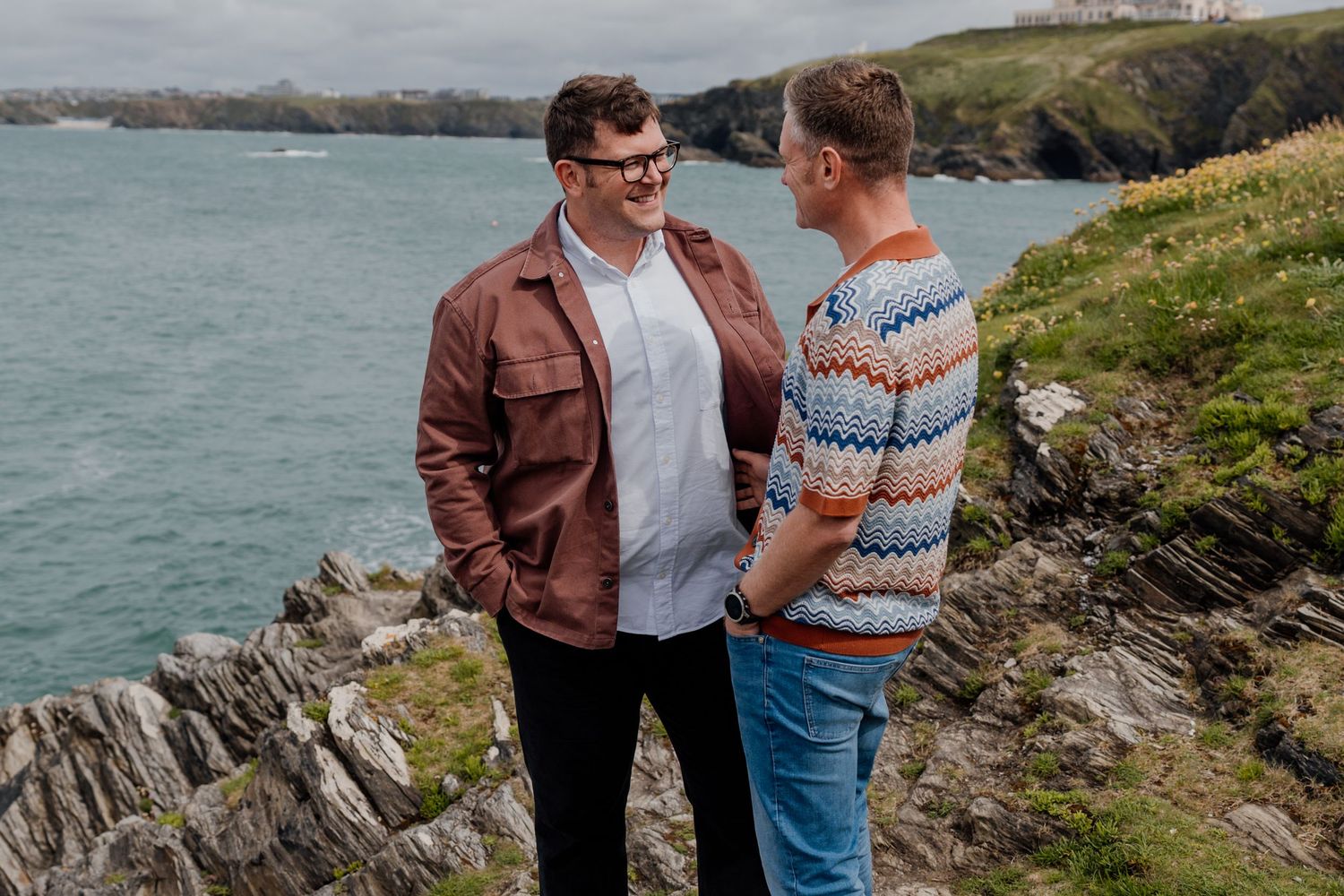 Two men stand on rocky coastal cliffs, smiling at each other with turquoise ocean waves in the background.