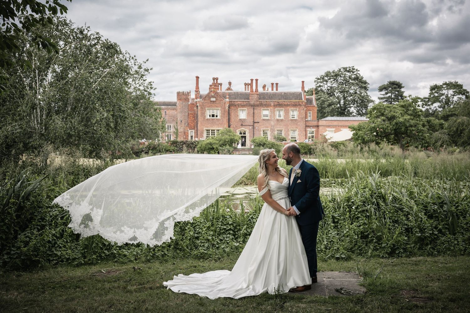Across the lake at Hodsock Priory, newlyweds embracing as the bride's veil blows in the wind.