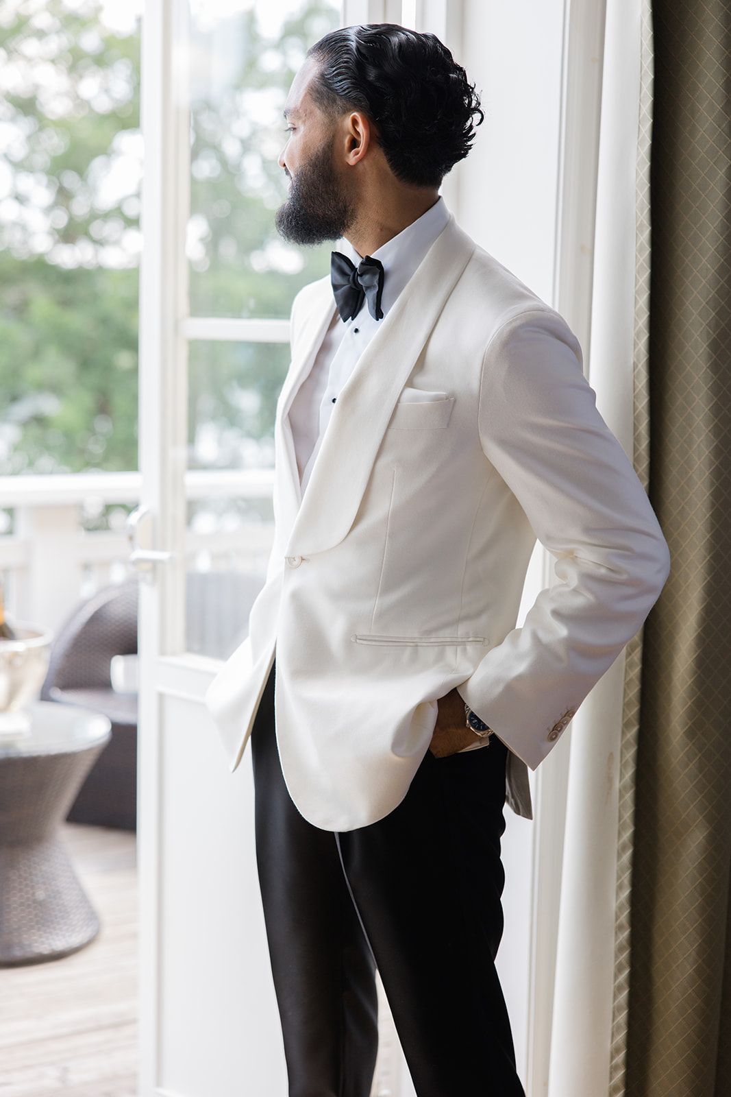Person in formal white tuxedo jacket and black pants poses in elegant interior setting near windows.