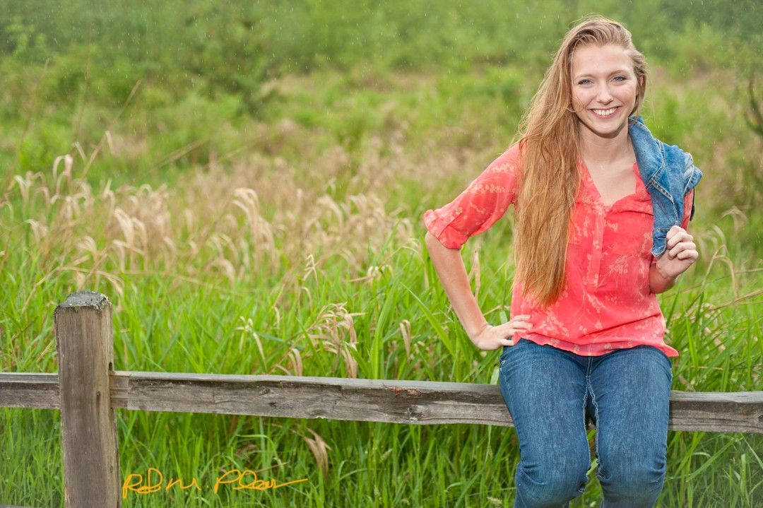 High school senior female at Nathan Chapman Memorial trail, Puyallup, sitting on fence with grassy field behind her.