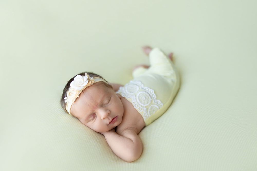 Sleeping newborn wearing white lace wrap and delicate headband rests peacefully on mint green backdrop.