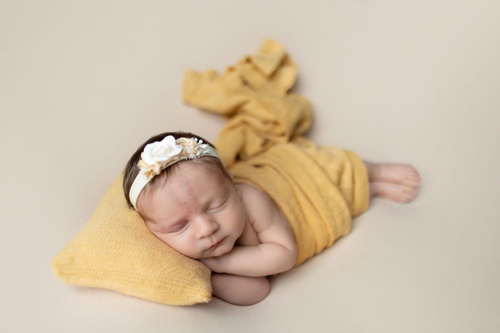Sleeping newborn wrapped in soft yellow fabric with matching decorative accessories on neutral backdrop.