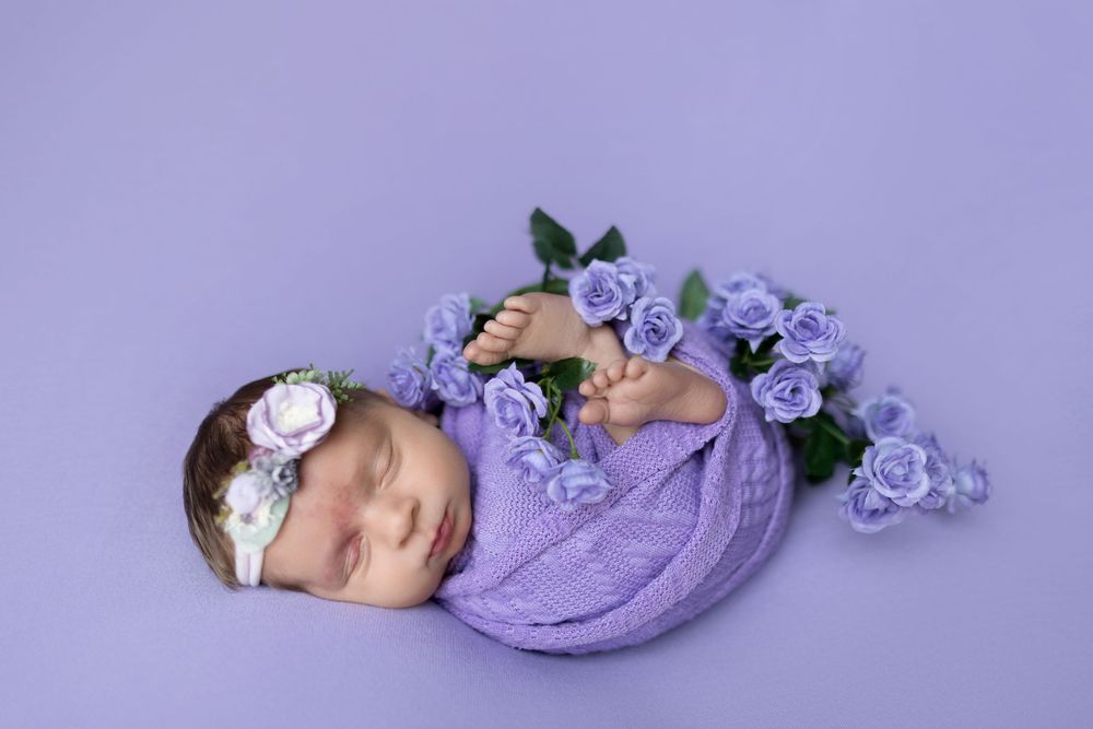 Sleeping newborn wrapped in purple swaddle with delicate lavender flowers on purple backdrop.