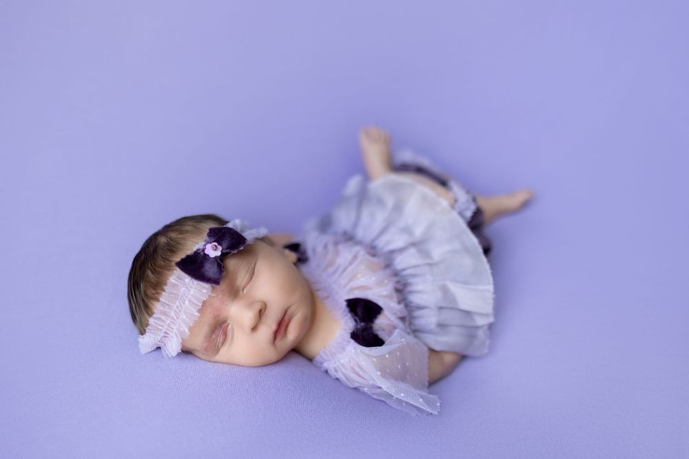 A baby wearing a white ruffled dress and bow headband lies on a purple backdrop during a photography session.