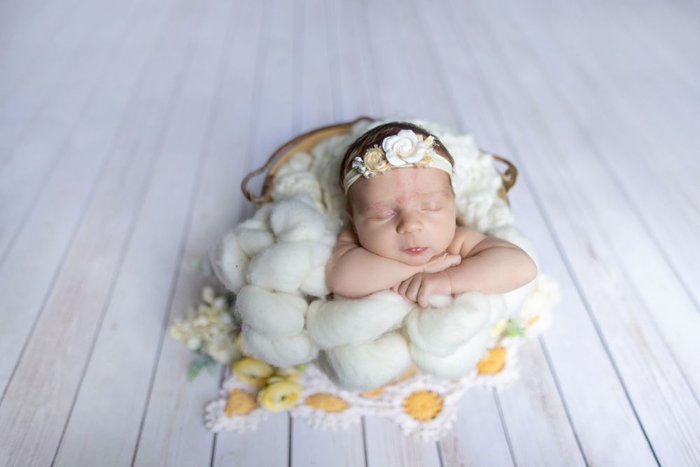 Sleeping newborn rests peacefully on soft white blanket surrounded by cotton balls and wearing delicate headband.