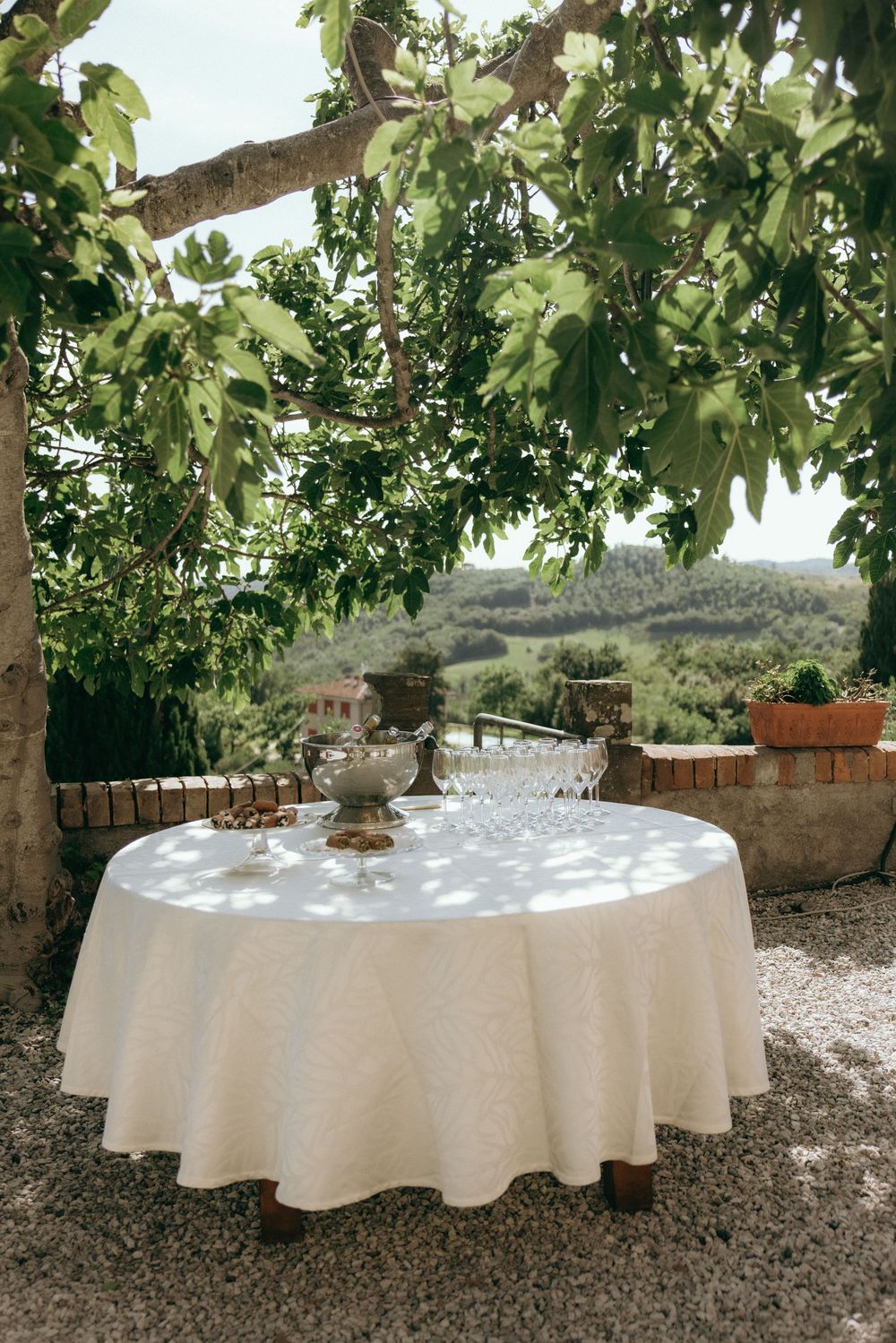 Round tables with white tablecloths set up under a leafy green canopy in an outdoor garden setting.