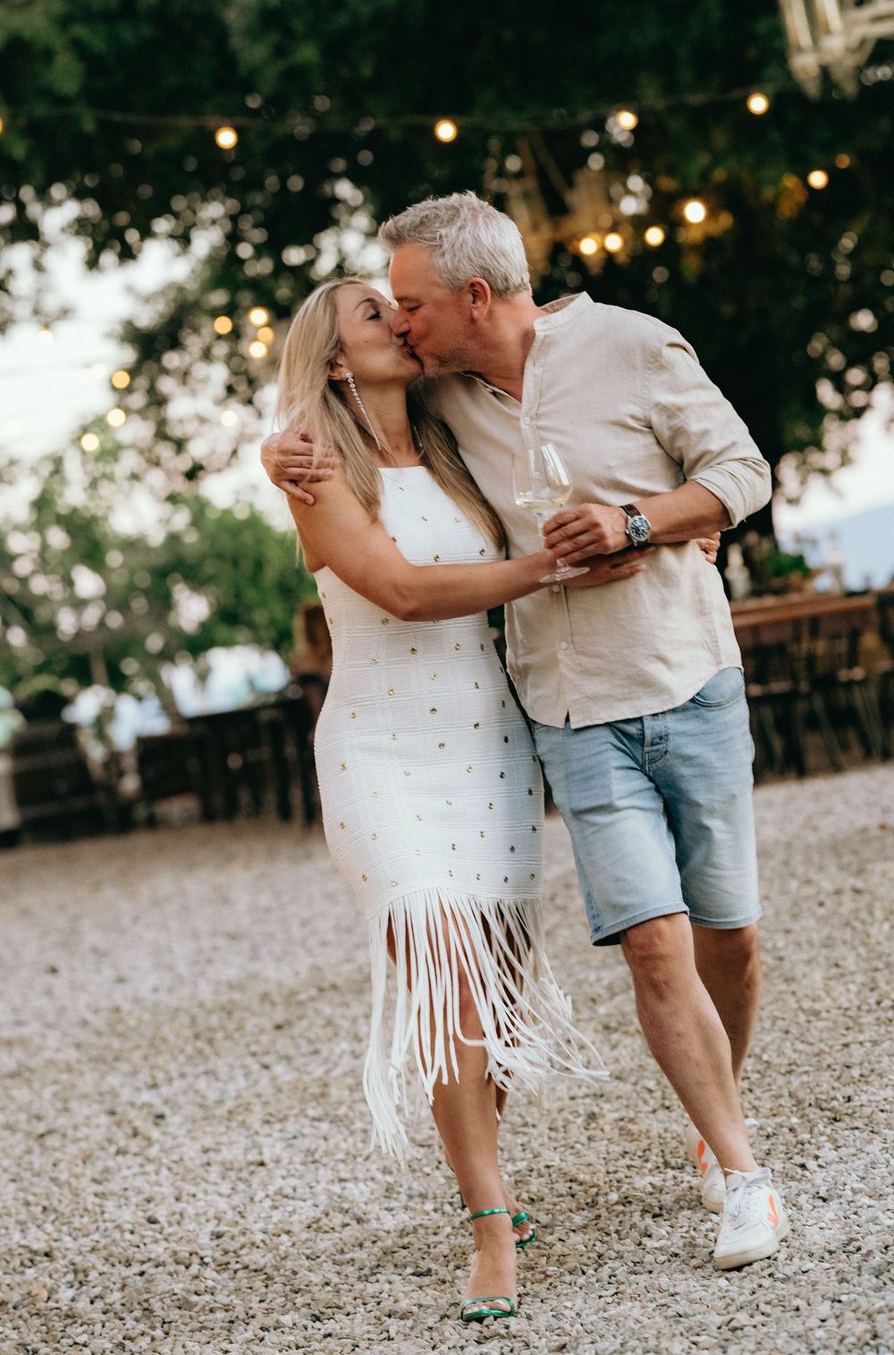 A romantic couple sharing an intimate moment on a gravel path with string lights overhead.