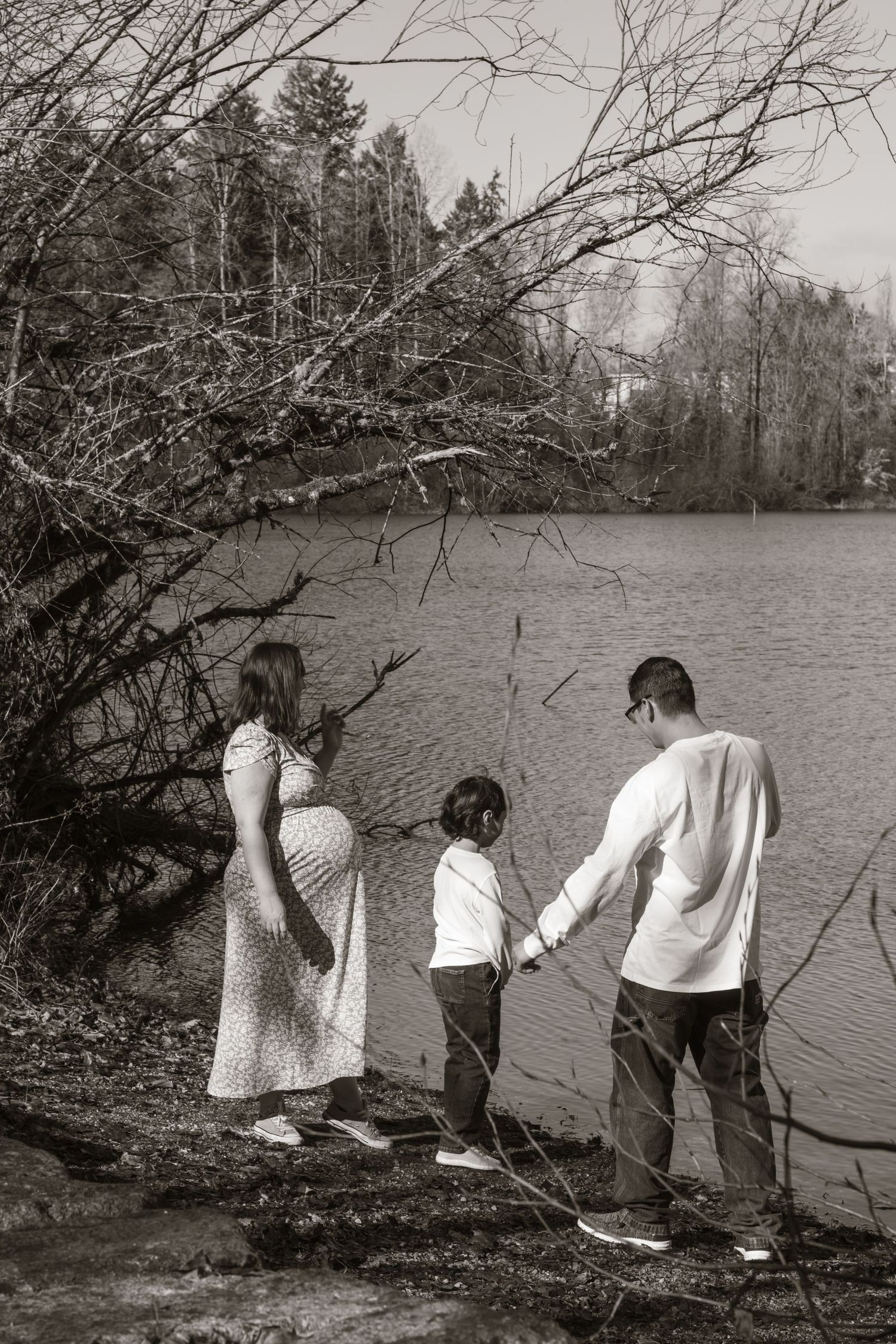 Black and white photo of pregnant woman, child, and man holding hands by a serene lakeside.