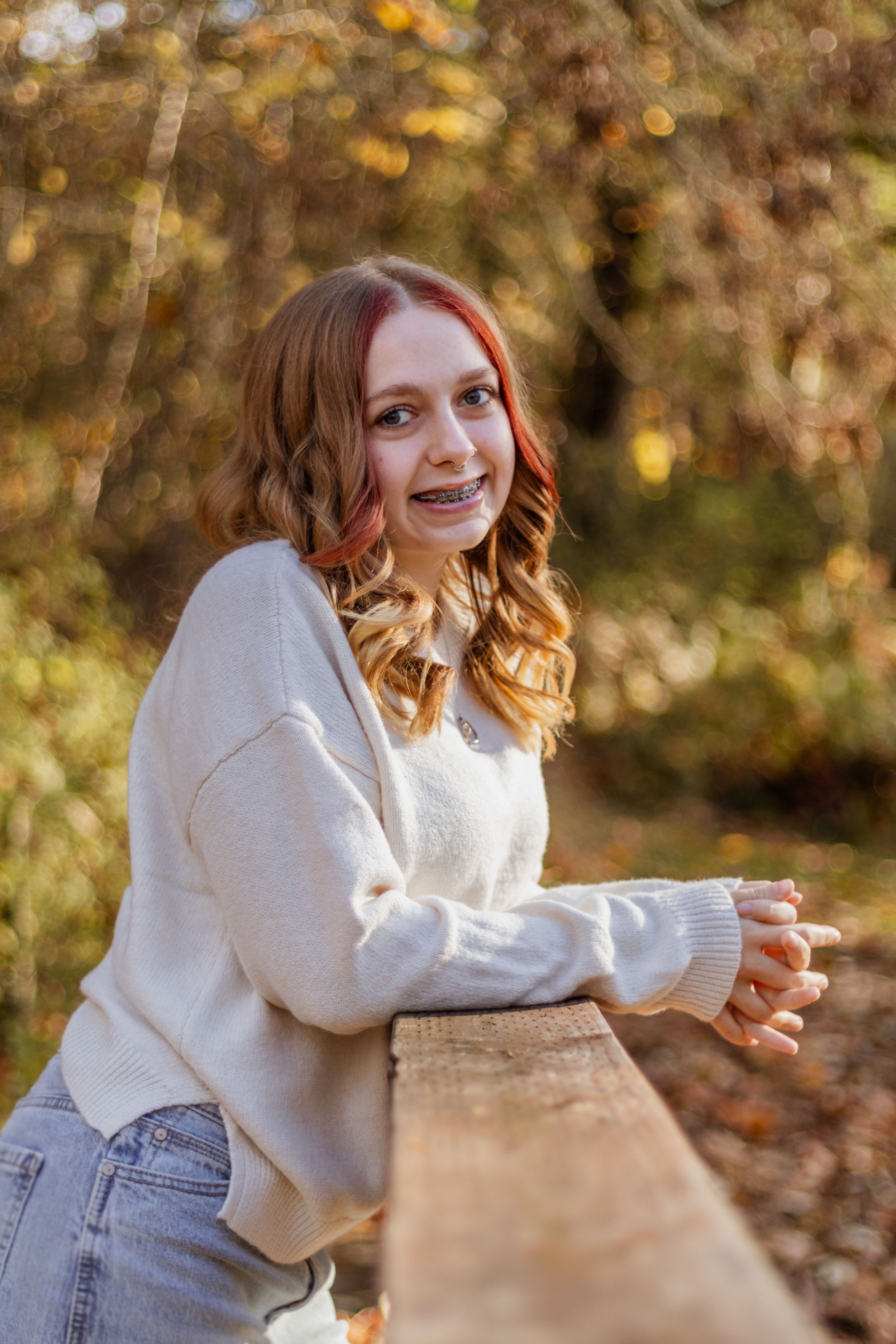 Smiling teenage girl with red hair and braces leans on wooden fence railing in autumn forest setting.