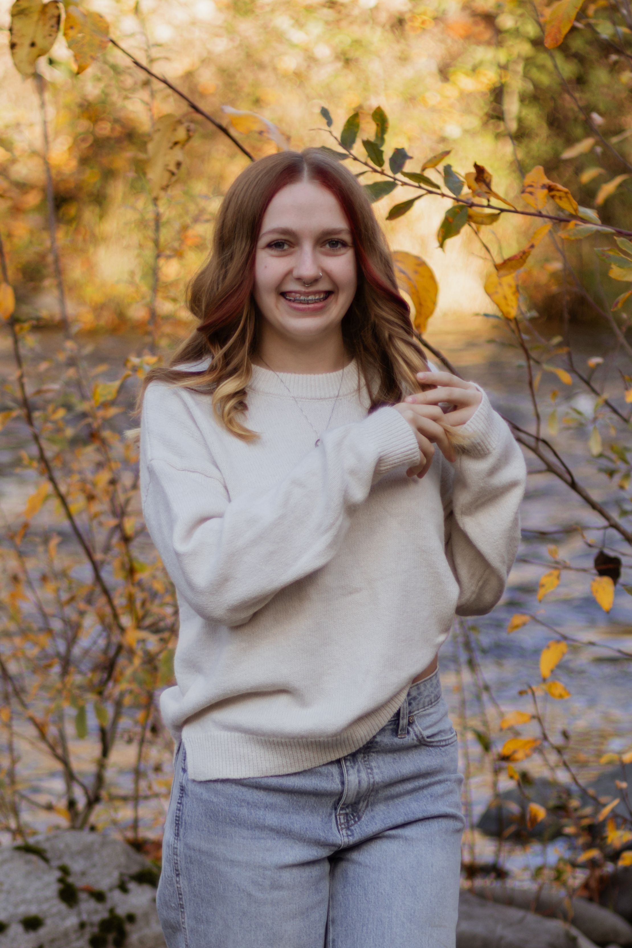 Smiling teenage girl with braces in cream sweater stands among golden autumn branches near a creek.