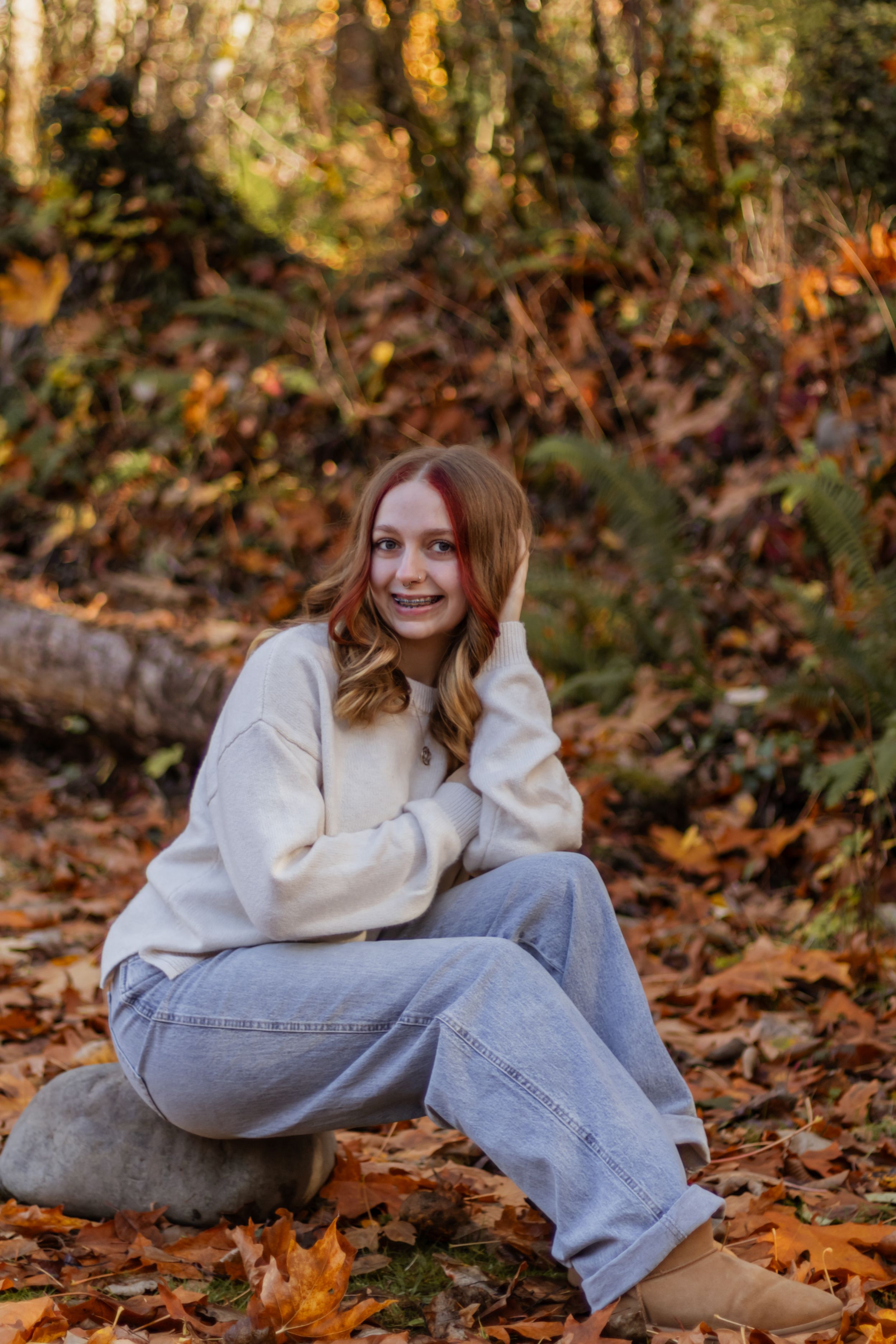 Teenage girl in cream sweater and jeans sits on rock surrounded by colorful fallen autumn leaves in forest.