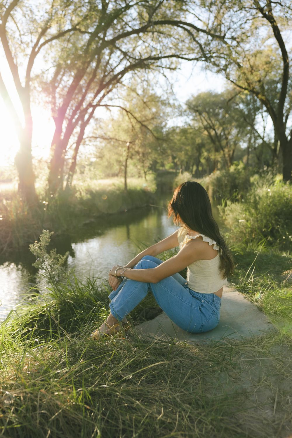 Person in blue dress relaxes on grassy riverbank during golden hour as sunlight filters through trees.