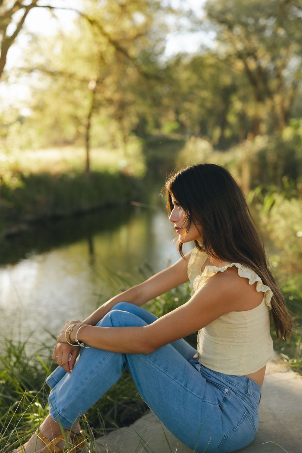 Person in blue jeans and white top sits thoughtfully by a serene lake at sunset wearing a protective face mask.