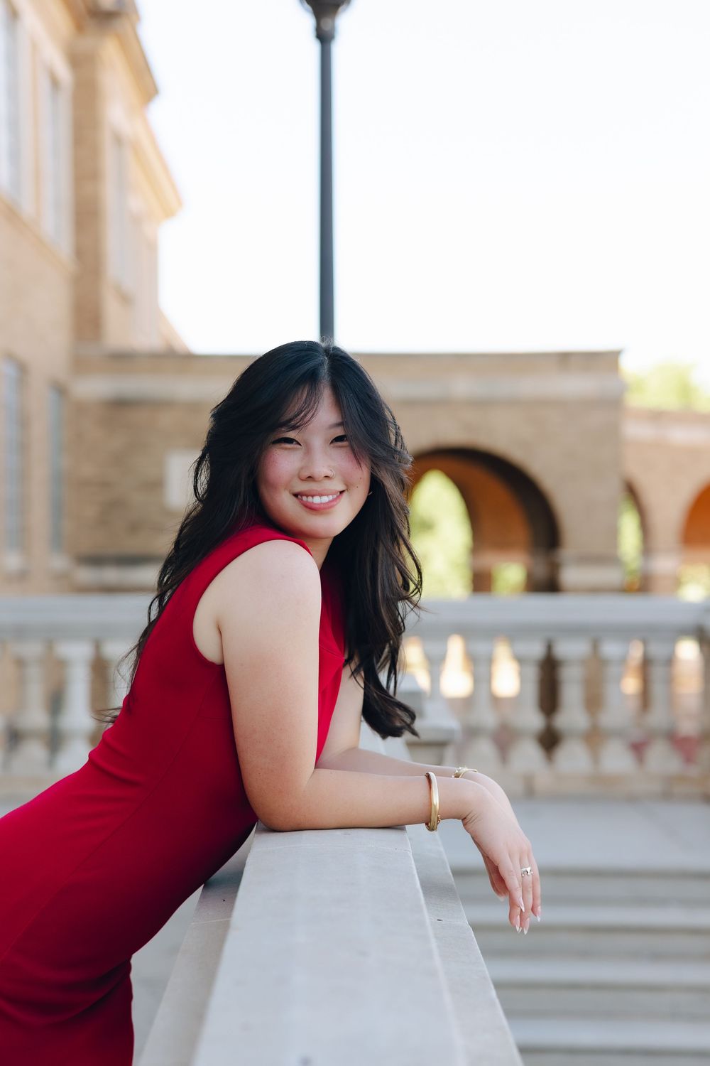 Woman in red sleeveless dress poses gracefully against stone columns in elegant outdoor architectural setting.