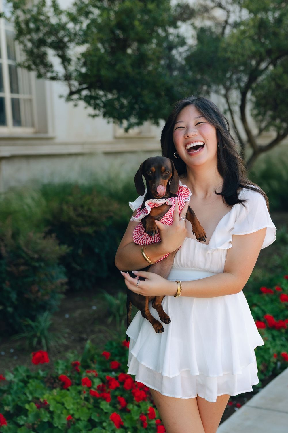 Woman in white dress laughing while holding a small dachshund puppy in a garden with red flowers.
