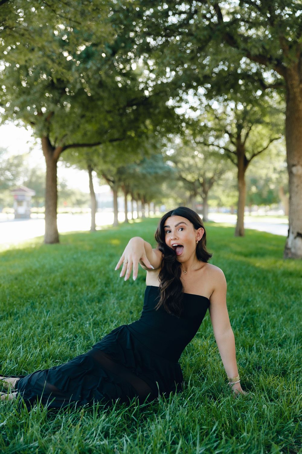 A person in a black dress poses playfully while sitting on green grass beneath a row of trees in a park setting.