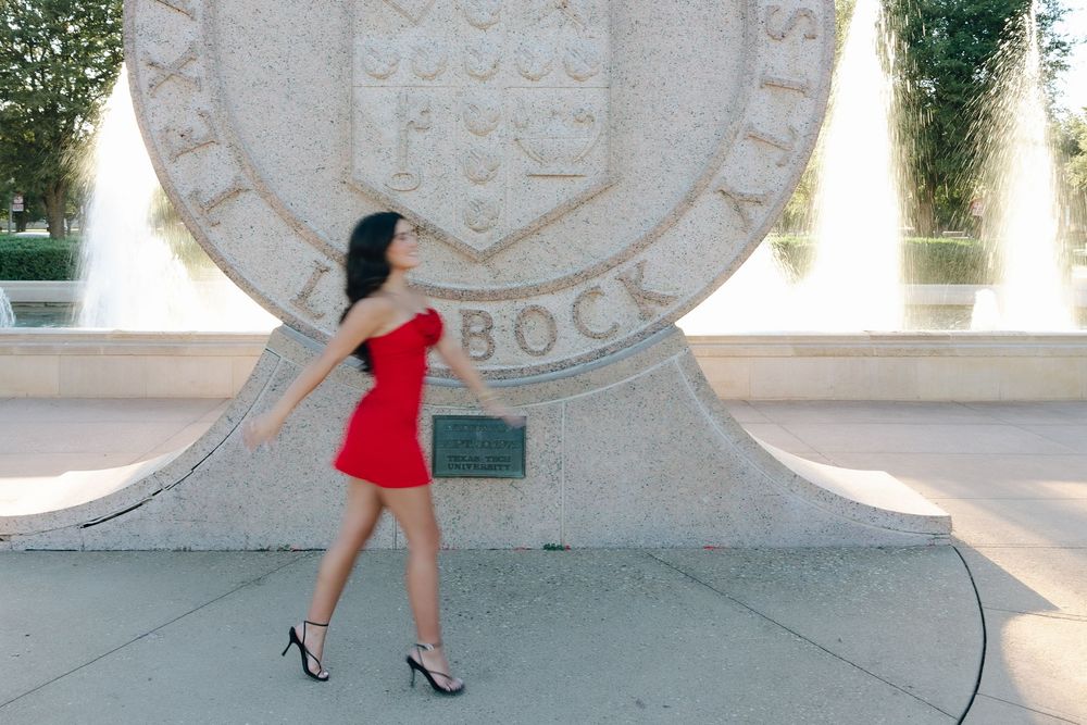 A person in a red dress and heels poses dramatically against a large stone monument or seal structure.
