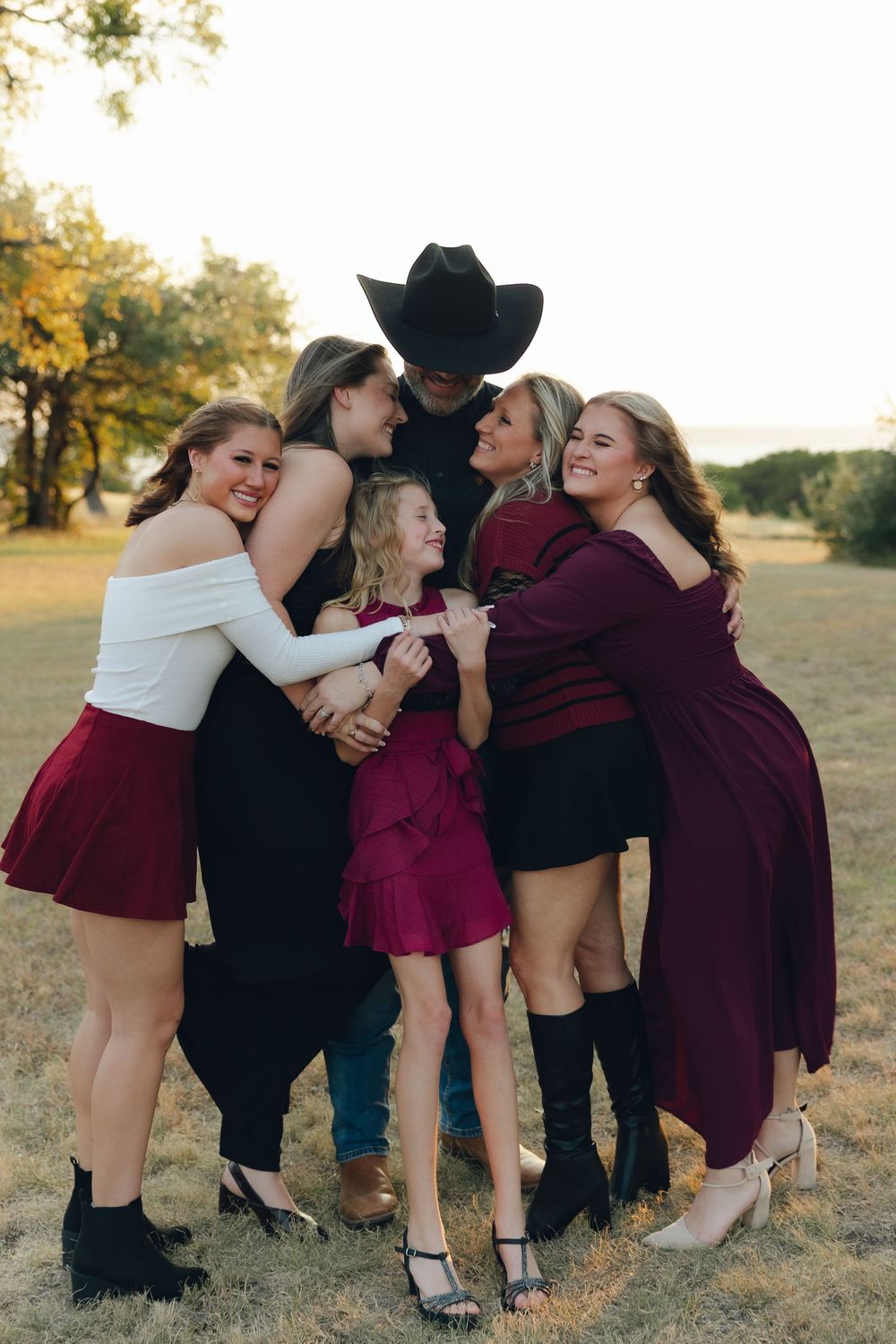 A group in burgundy dresses and black attire share joyful candid moments in a grassy outdoor setting at sunset.