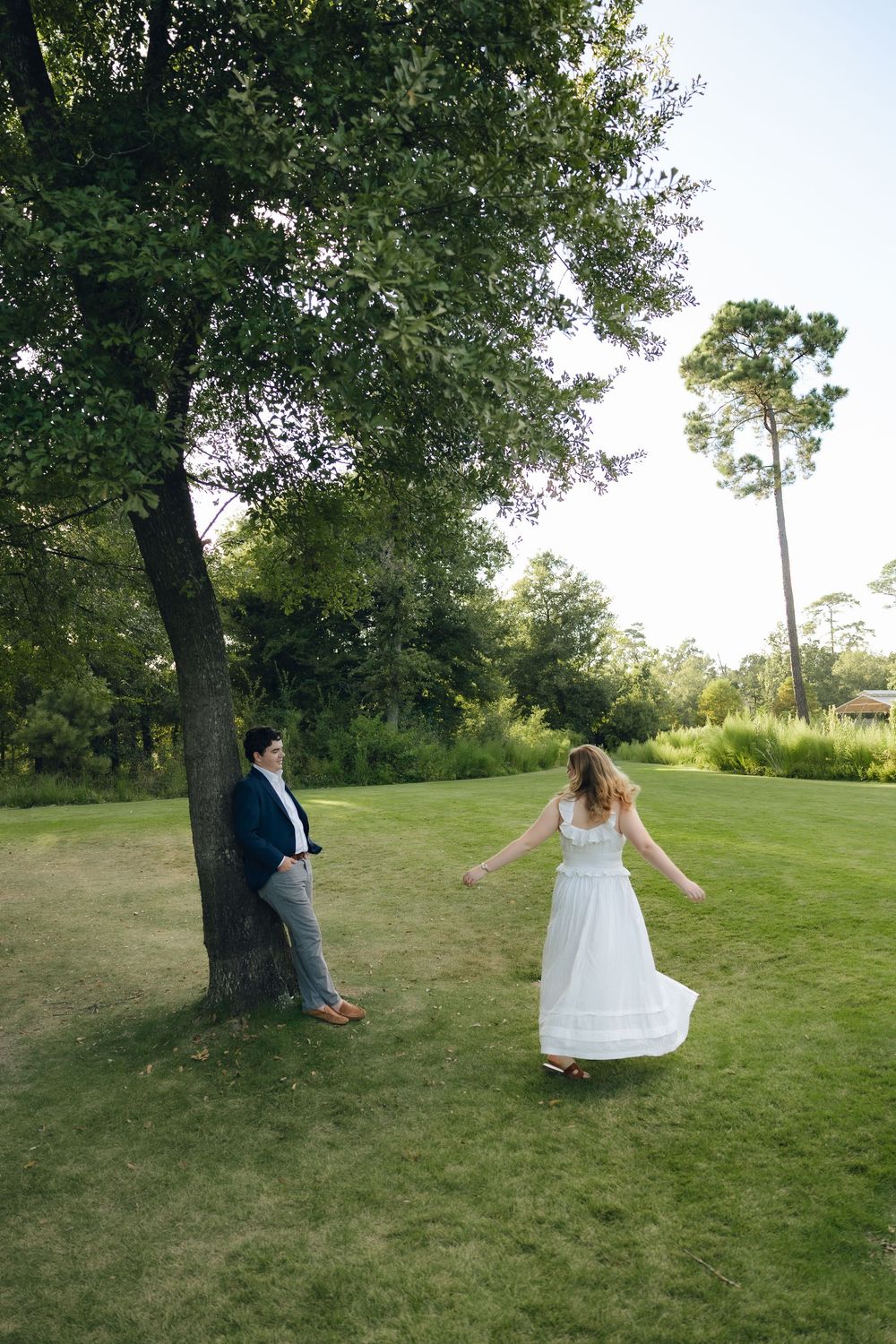A couple in wedding attire poses playfully by a large tree on a grassy lawn during a summer photoshoot.