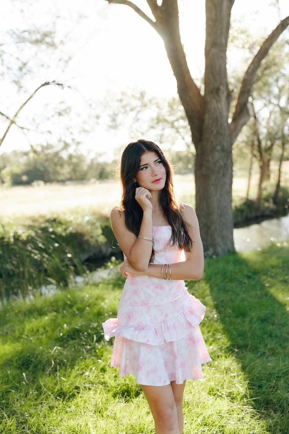 A person in a pink and white floral dress poses gracefully in a sunlit outdoor setting with trees in the background.