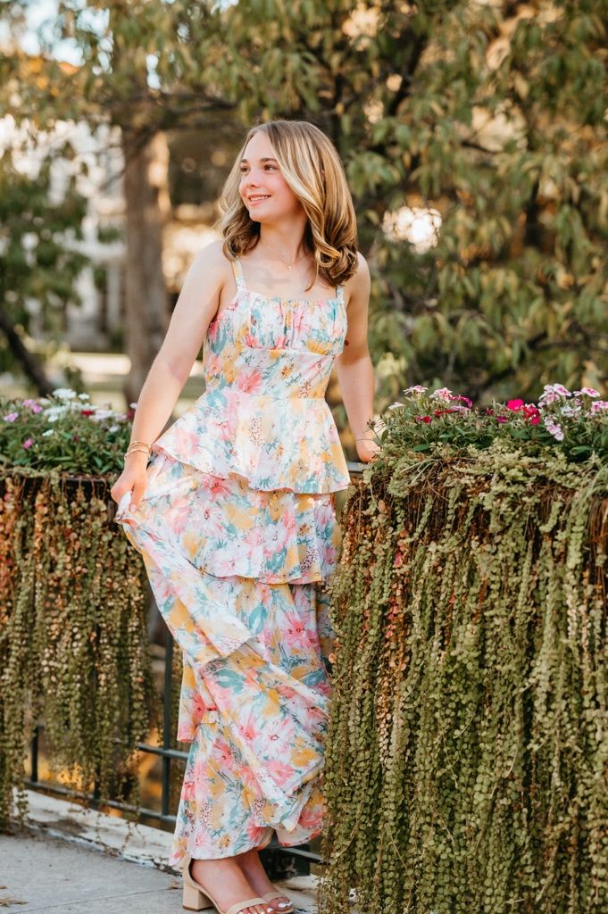 A person wearing a pastel floral dress poses near decorative hanging plants and greenery outdoors.
