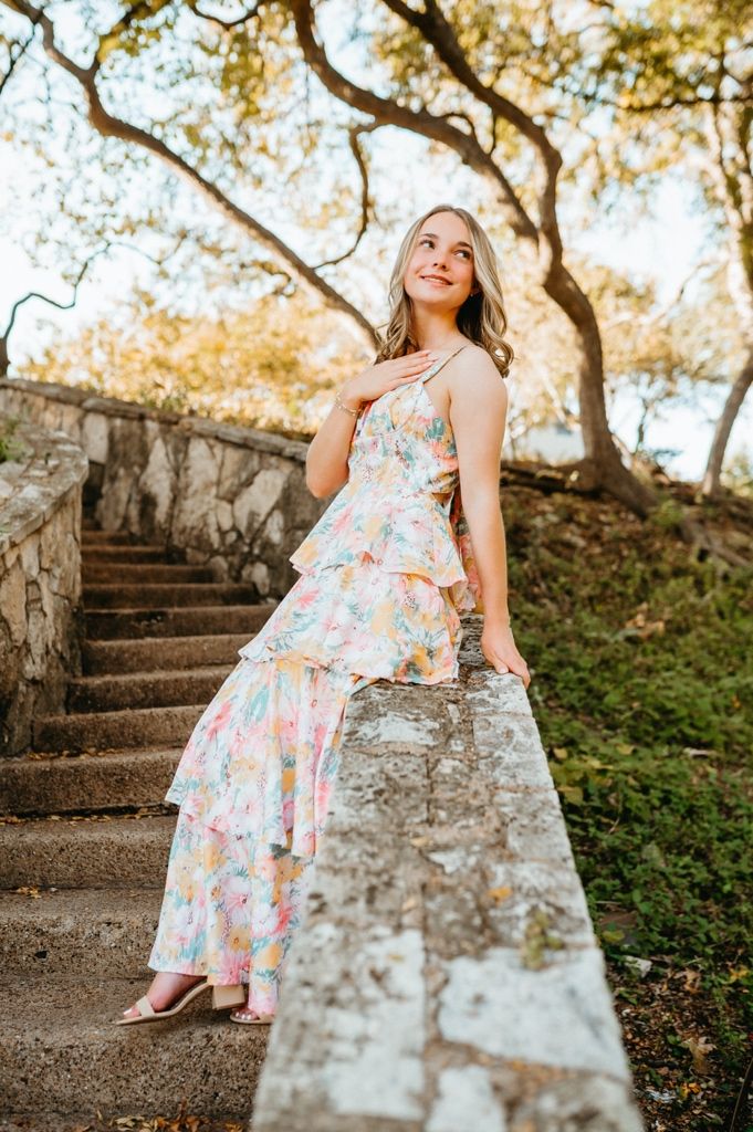 A woman in a tiered floral maxi dress stands on stone steps surrounded by autumn trees and dappled sunlight.