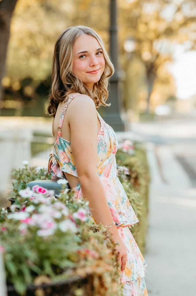 A young woman in a floral summer dress poses outdoors with soft golden lighting and flowers in the background.