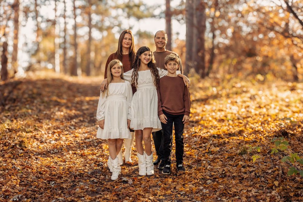 A group stands together on fallen autumn leaves in a sunlit wooded area wearing coordinated cream and brown outfits.