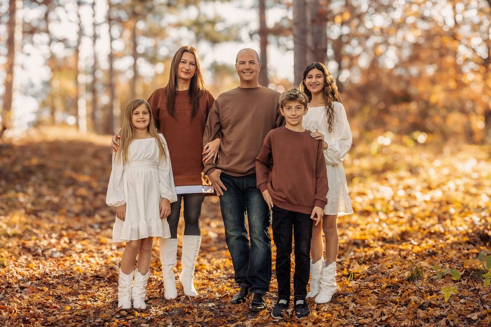 A family group stands together outdoors in autumn with fallen leaves and trees in the background.