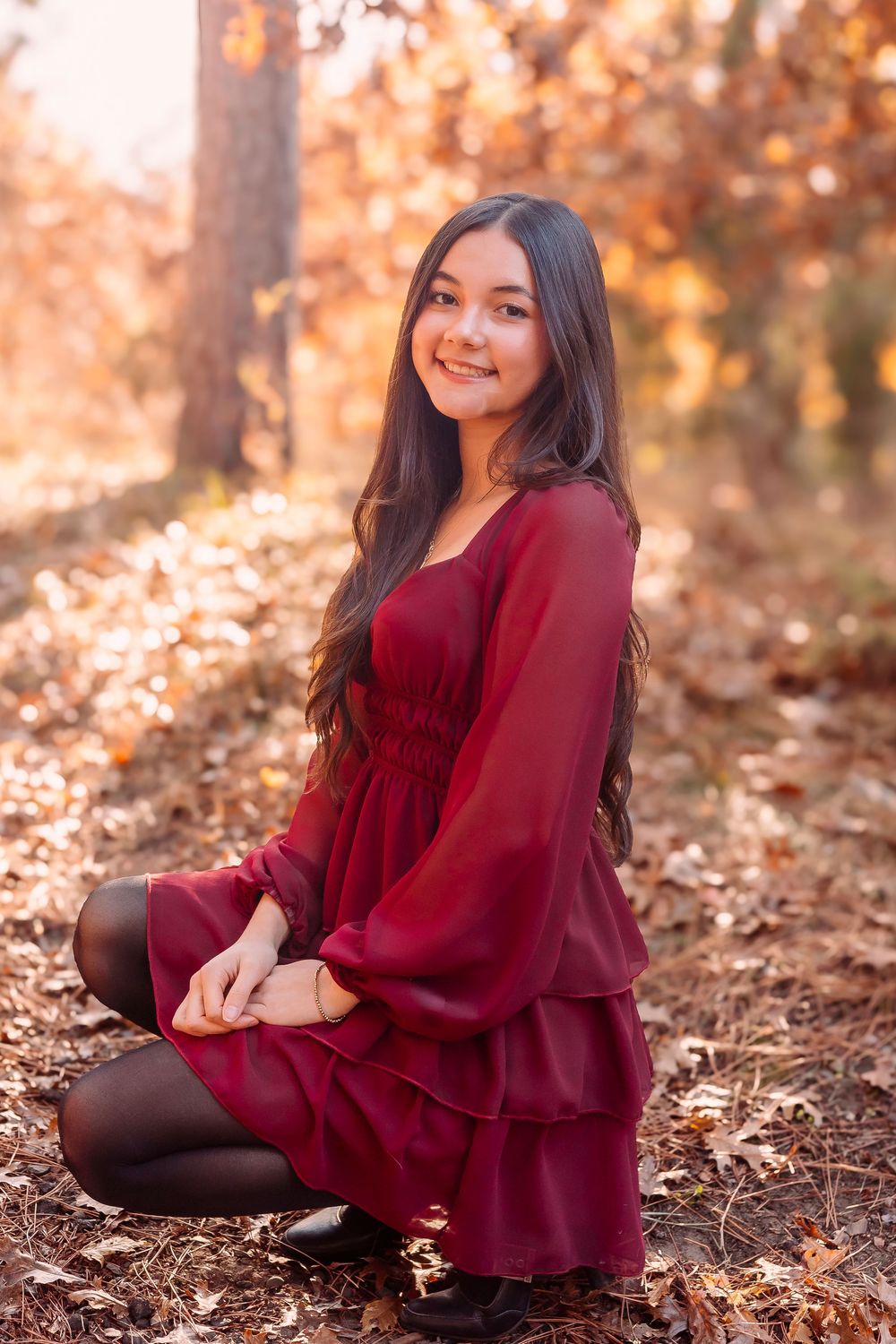 A person in a burgundy dress poses outdoors among fallen autumn leaves on a sunny fall day.