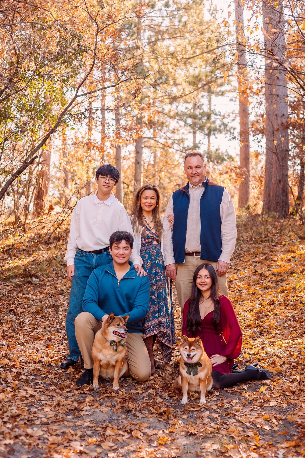 Group portrait with two dogs sitting among fallen autumn leaves in a wooded setting during golden hour.