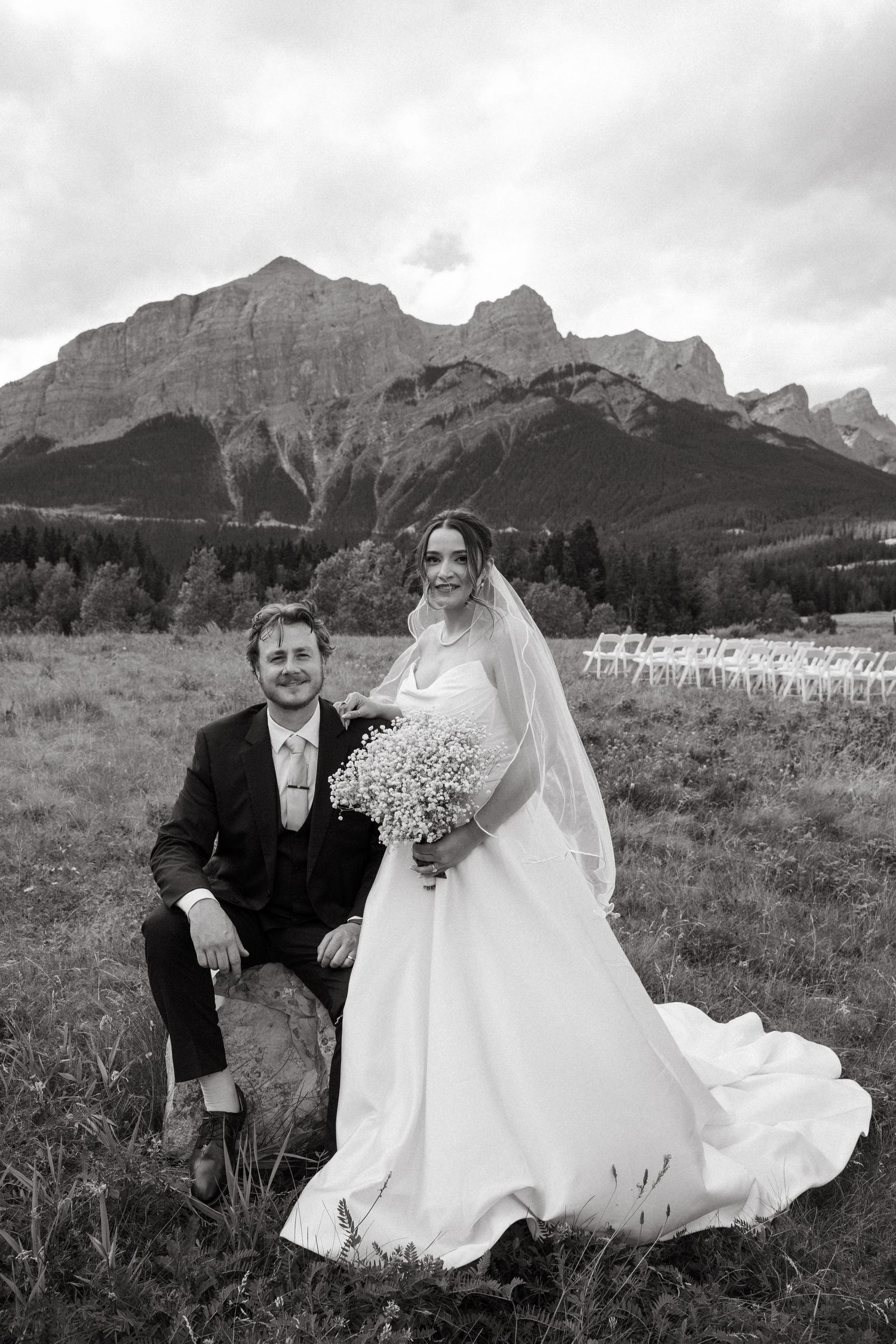 A wedding couple sits together in a grassy meadow with dramatic mountain peaks rising in the background under cloudy skies.