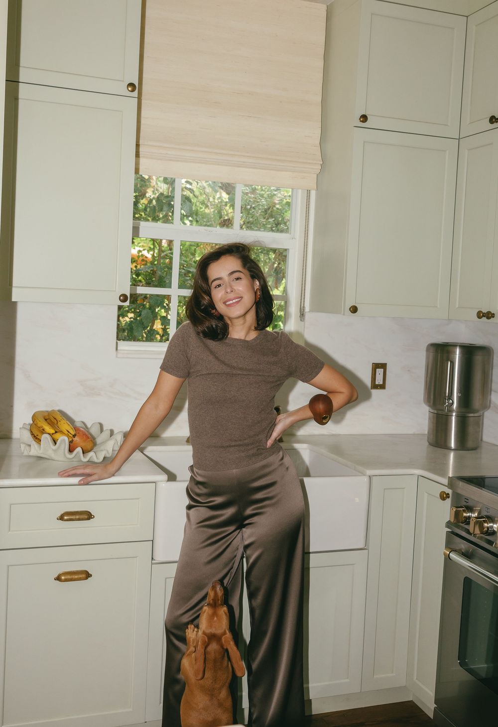 Woman in brown top and satin pants smiling in a stylish white kitchen with marble countertops and green cabinets.