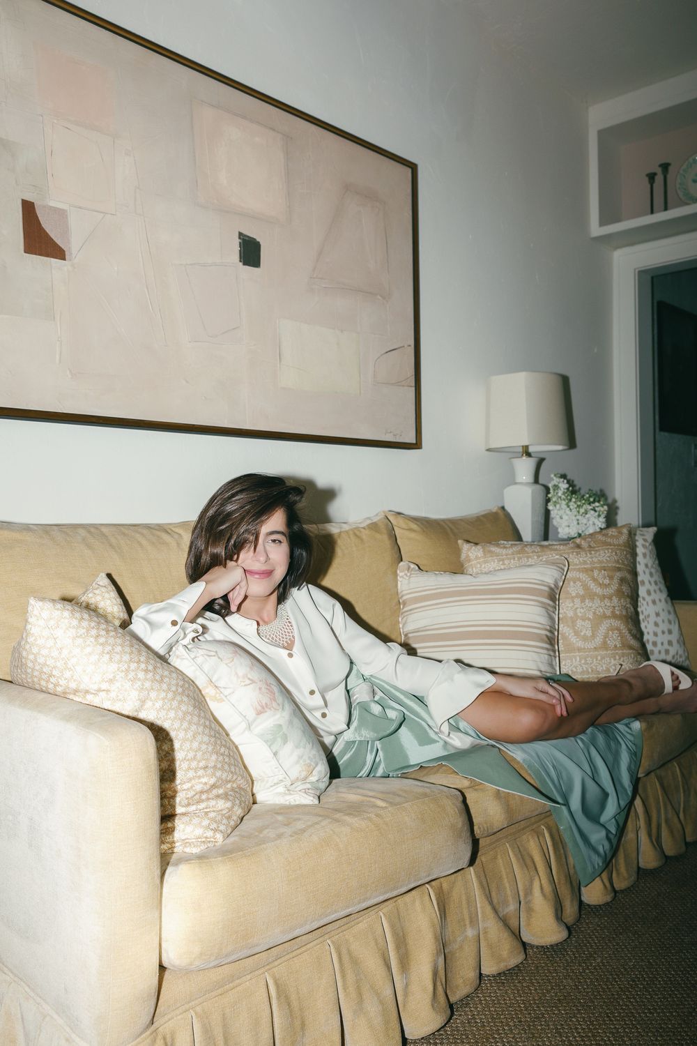 Woman in white top and mint skirt relaxing on a beige skirted sofa with decorative pillows and abstract art.