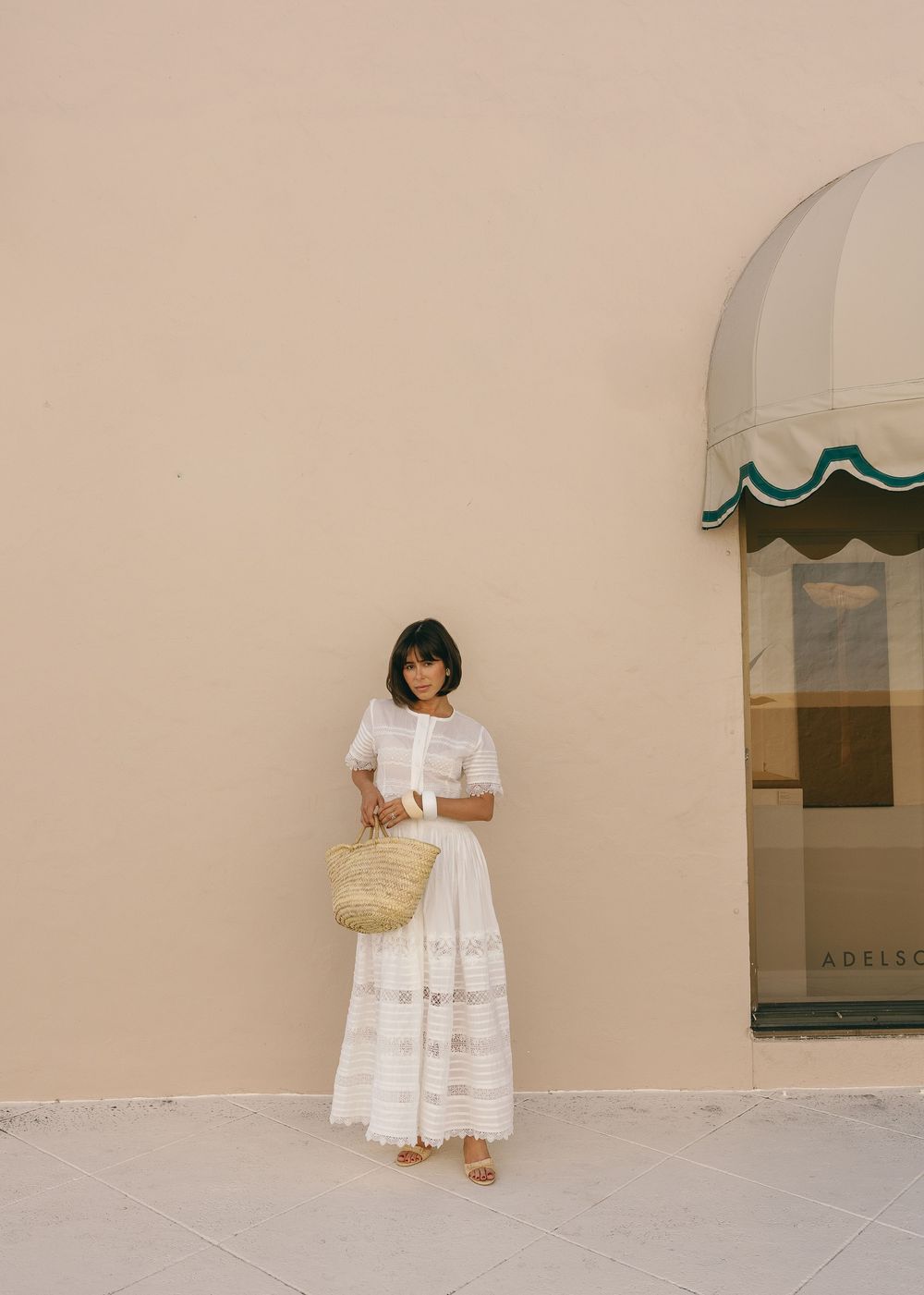 Woman in white lace midi dress holding a straw bag, standing against a beige wall near a striped awning.