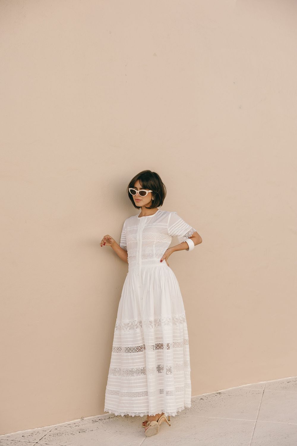 Woman in white lace maxi dress and sunglasses posing against a beige wall in elegant summer style.