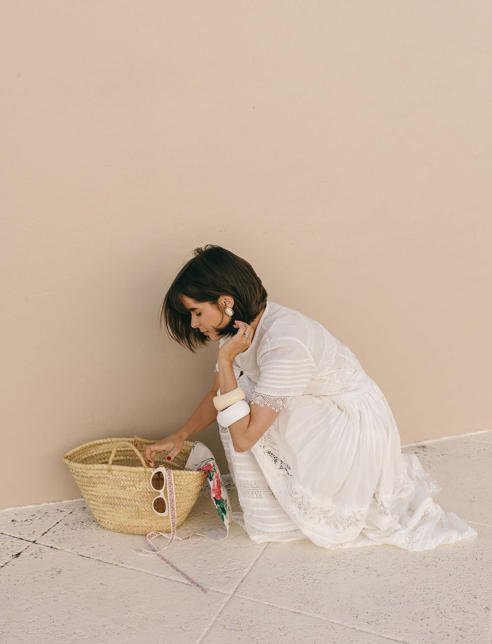 Woman in white dress kneeling on tile floor, reaching into a straw tote bag with sunglasses beside a beige wall.