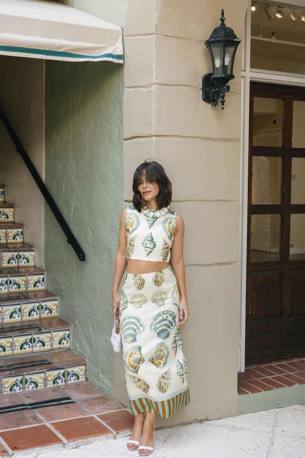 Woman in white seashell print crop top and midi skirt standing by building entrance with tiled steps.