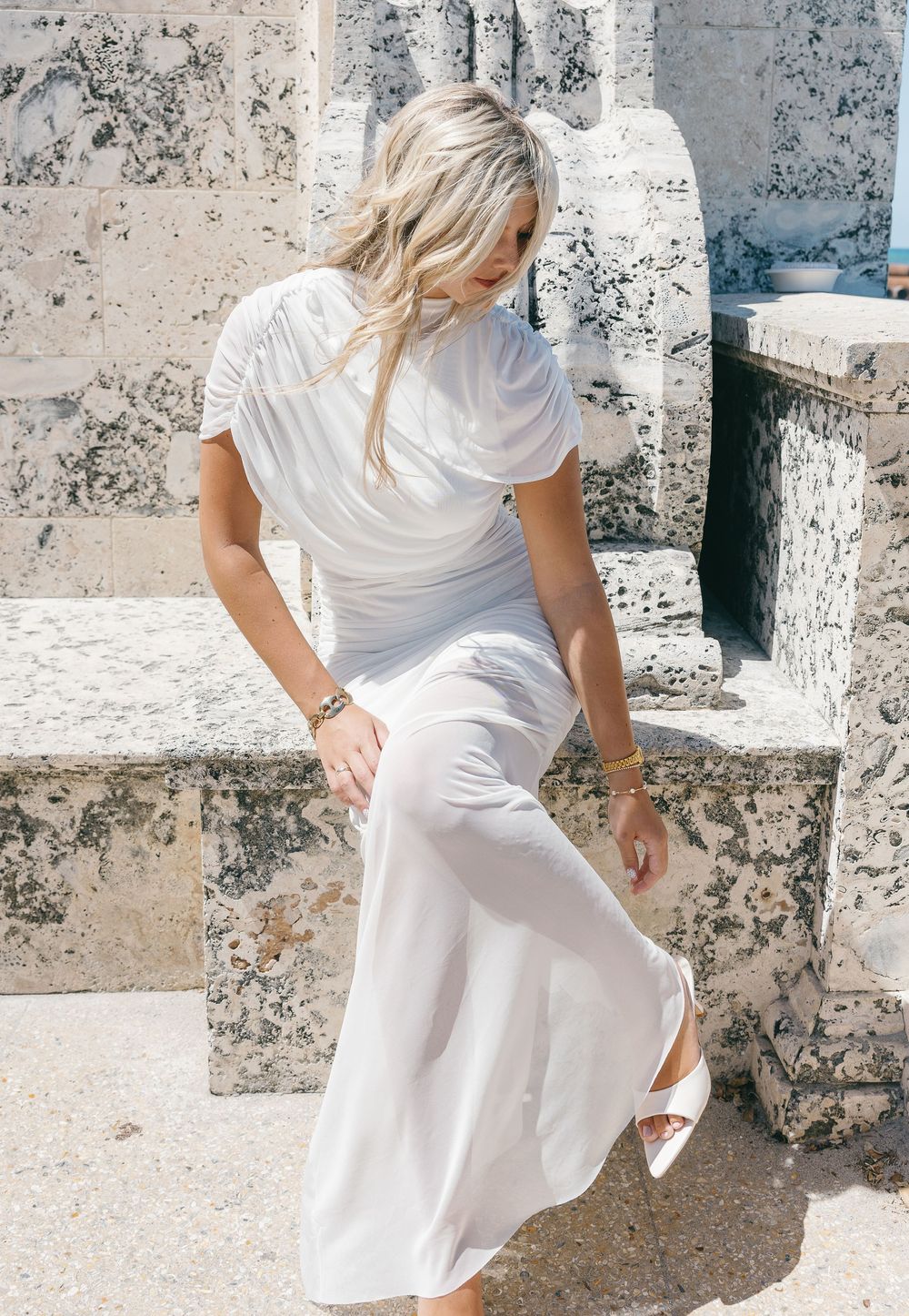 Blonde woman in elegant white ruched midi dress and white heels sitting on stone steps outdoors.