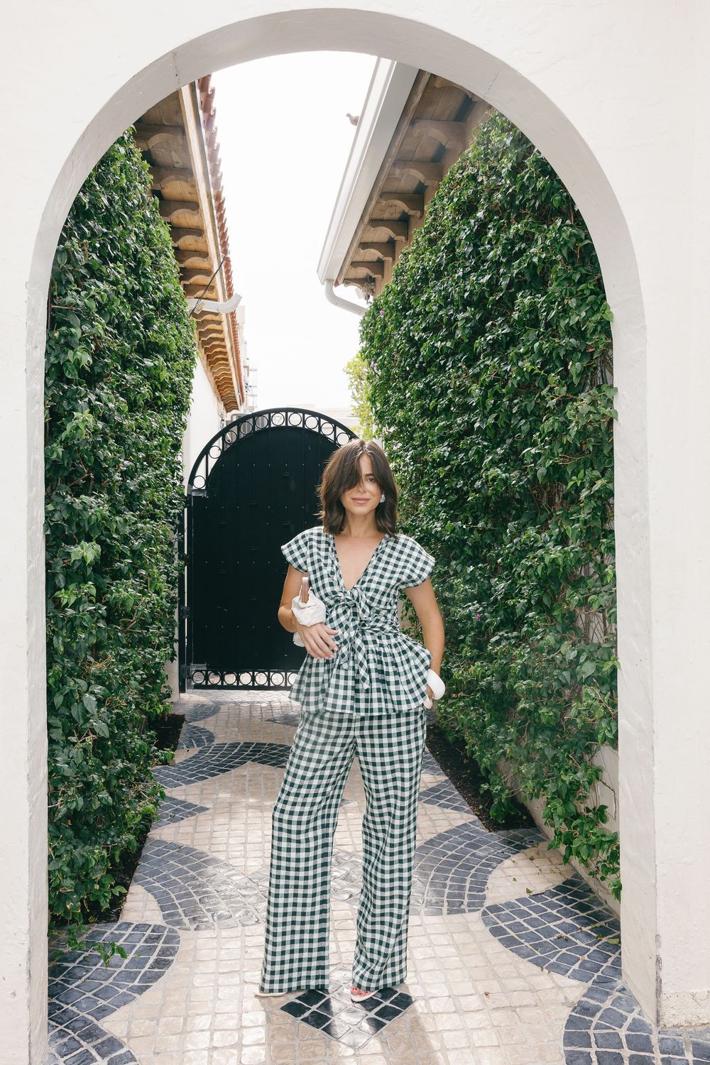 Woman in green gingham matching set poses under white arch surrounded by lush ivy walls on decorative tile path.