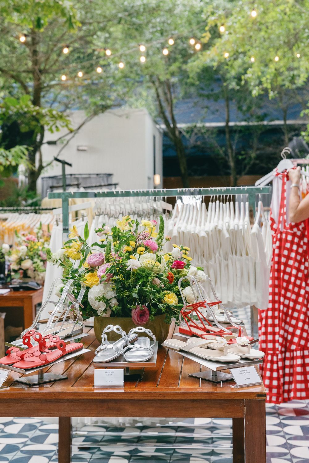 Outdoor boutique display with sandals, floral arrangement, white hanging garments, and a red gingham dress under string lights.