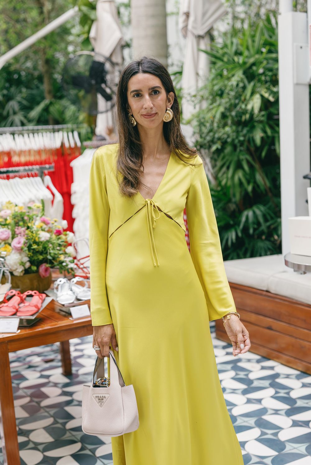 Woman wearing a yellow-green long sleeve maxi dress holding a white Prada bag, standing in a lush tropical outdoor setting.