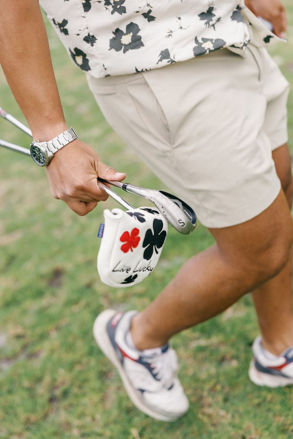 Golfer holding a putter with a Lucky Clover headcover on a golf course, wearing khaki shorts and a floral shirt.