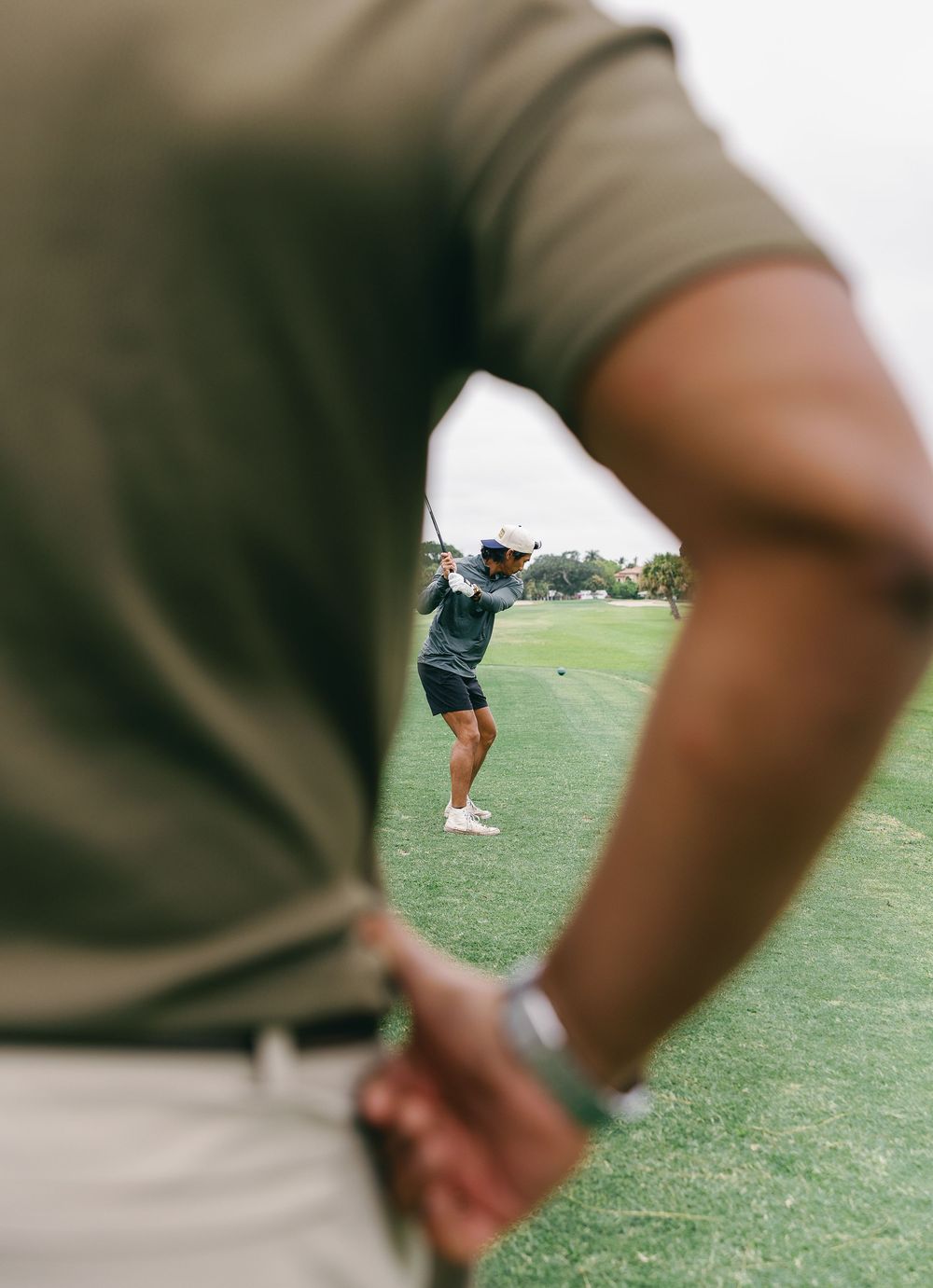 Golfer completing a swing on a sunny course, viewed over the shoulder of another player watching nearby.