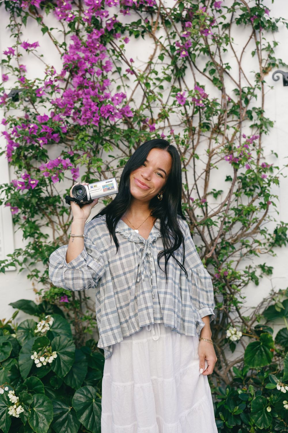 Young woman in plaid top holding a video camera, posing in front of a white wall with purple bougainvillea flowers.