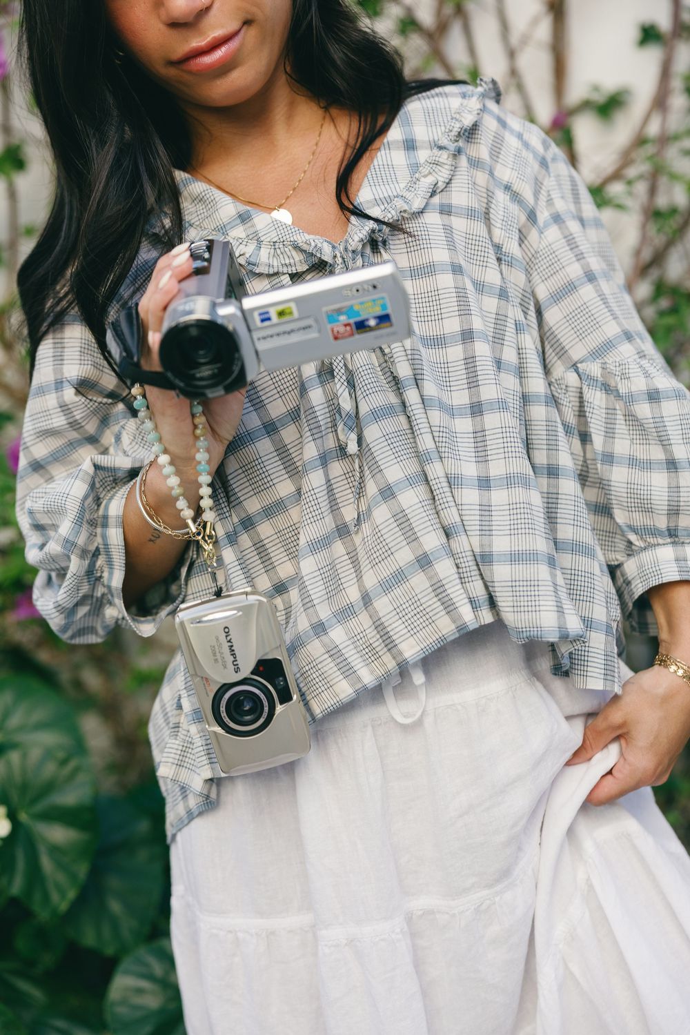 Woman in plaid blouse holding vintage camcorder with mini camera bag accessory, standing outdoors.