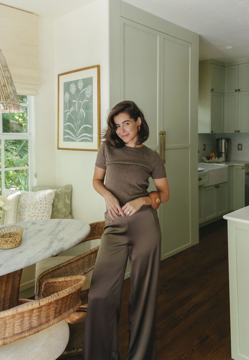Woman in brown top and wide-leg pants standing in a sage green kitchen with marble dining nook.